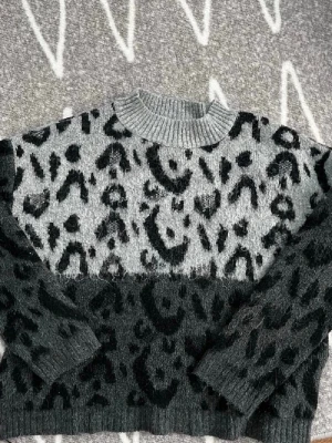 Grå och svart leopardmönstrad tröja - Stickad tröja med leopardmönster i grått och svart. Tröjan har rund halsringning, långa ärmar och en mjuk, fluffig känsla. Perfekt för dig som gillar djurmönster och vill ha en trendig look.