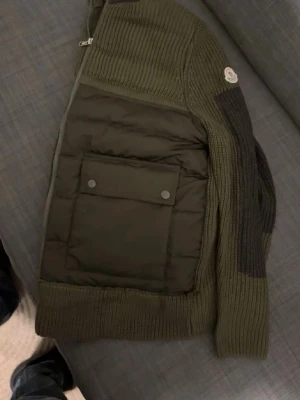 Moncler cardigan  - Snygg olivgrön cardiganjacka från Moncler med quiltad front och stickade partier. Jackan har två stora fickor med tryckknappar, ribbade muddar och detaljer i mörkgrått på ärmar och krage. Dragkedja framtill och Moncler-logga på ärmen.