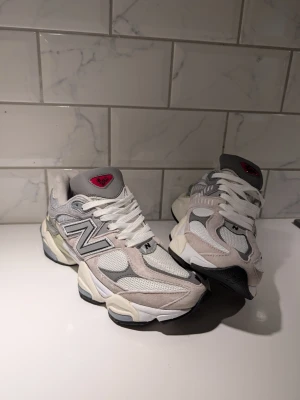 New Balance grå/vita sneakers - Snygga sneakers från New Balance i grått, vitt och beige med mesh och mockadetaljer. Klassisk N-logga på sidan och coola detaljer på sulan. Skorna har vita skosnören och en röd detalj på plösen. Perfekta för dig som gillar streetwear och sportig stil.