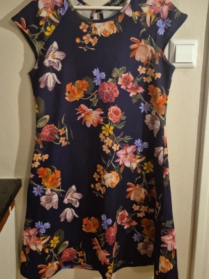Mörkblå blommig knälång klänning - Knälängd klänning i mörkblått med färgglatt blommönster i rosa, orange, gult och lila. Klänningen har rund halsringning och är ärmlös, perfekt för en somrig look. Materialet känns mjukt och följsamt, polyester.