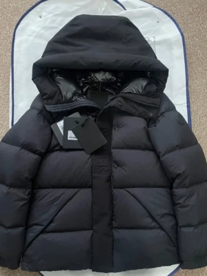 Svart pufferjacka från Moncler - Säljer en svart pufferjacka från Moncler med huva och diskret logga på bröstet. Jackan har breda, vadderade paneler, tryckknappar och dragkedja framtill. Perfekt för kalla vinterdagar och riktigt snygg till streetwear-looken.