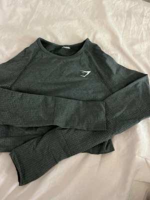 Mörkgrå långärmad crop top Gymshark - Sportig mörkgrå långärmad crop top från Gymshark med diskret mönstrad struktur och logga på bröstet. Tillverkad i ett stretchigt och ventilerande material som passar perfekt till träning och gym. Tight passform och rund hals. Vet inte storlek med passar mig som har xs/s