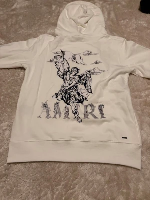 Vit amiri hoodie - Säljer en vit hoodie från Amiri med stor grafisk ängel och text på ryggen samt mindre tryck på bröstet. Hoodien har huva och en klassisk magficka framtill. Tillverkad i mjukt material, perfekt för en clean och stilren streetwear-look.