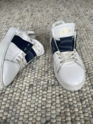 Valentino vita sneakers med svart detalj - Snygga vita sneakers från Valentino Garavani med bred svart läderdetalj över sidan och klassisk logga på plösen. Skorna har vita snören och en chunky sula som ger en modern vibe. Perfekta för dig som vill ha en clean men ändå statement look.