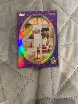 Topps Real Madrid samlarkort Pepe - Samlarkort från Topps med Real Madrid-tema, föreställande spelaren Pepe. Kortet har lila bakgrund med gulddetaljer och holografisk effekt, samt numrering 243/250. Baksidan visar att Pepe vunnit 14 troféer. Perfekt för samlare!