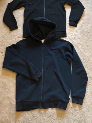 2 x  hoodie Jack & Jones strl S - Klassisk svart hoodie från Jack & Jones med dragkedja framtill och stor huva. Hoodien har ribbade muddar vid ärmslut och nederkant samt en känguruficka. Tillverkad i mjukt bomullsmaterial som är skönt mot huden. Perfekt för en avslappnad och stilren look.