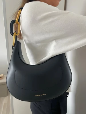 Svart axelväska från Charles & Keith - Snygg svart axelväska i halvmåneform från Charles & Keith. Väskan har stora guldfärgade och svarta länkar på axelremmen och är gjord i slätt konstläder. Minimalistisk design med diskret logga framtill. Perfekt för dig som gillar trendiga accessoarer.