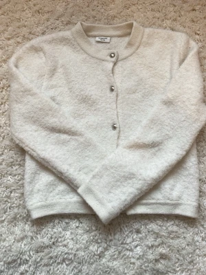 Vit fluffig kofta från Kappahl - Mysig vit kofta från Kappahl med rund halsringning och silverfärgade knappar framtill. Koftan har en fluffig, mjuk textur och långa ärmar. Perfekt för lager på lager och enkel att matcha med olika outfits. Tröjan saknar en knapp längst nere