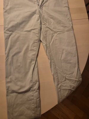 Beige chinos Asket - Snygga beige chinos med rak passform, tillverkade i 98% bomull och 2% elastan för extra komfort. Klassisk design med dragkedjegylf och bälteshällor. Perfekta för en avslappnad men stilren look.