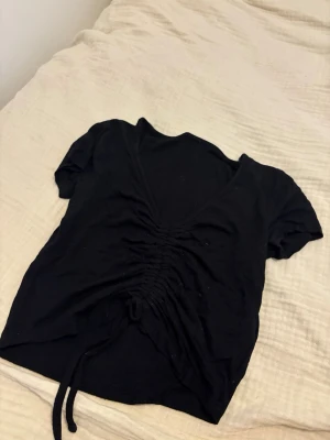 Svart topp med snörning och v-ringning - Snygg svart topp med korta ärmar och djup v-ringning. Toppen har en trendig snörning framtill som ger en rynkad effekt och kan justeras för önskad passform. Perfekt till jeans eller kjol för en cool look.