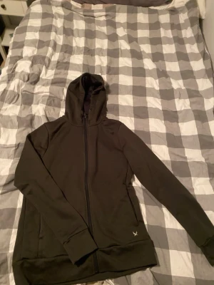 Grön hoodie med dragkedja från SOC - Svart hoodie från SOC med hel dragkedja och huva. Tillverkad i mjukt, stretchigt material och har två sidofickor samt diskret logga på framsidan. Perfekt för träning eller chill, enkel och stilren design.