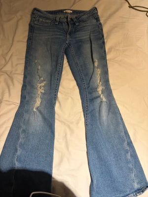 Blå bootcut jeans med slitningar - Säljer ett par ljusblå lågmidjade bootcut jeans från Gina Tricot 14+ med slitna detaljer på båda benen. Jeansen har klassisk femficksmodell, bälteshällor och dragkedja. 