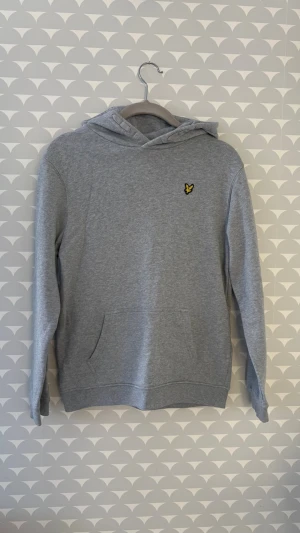 Grå hoodie från Lyle & Scott - Snygg grå hoodie från Lyle & Scott med klassisk gul logga på bröstet. Hoodien har huva, känguruficka och ribbade muddar vid ärmslut och nederkant. Tillverkad i mjukt bomullsmaterial som är perfekt för en avslappnad stil. Nypris 599kr💕