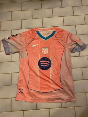 Rosa FC Barcelona matchtröja Nike - Säljer en rosa FC Barcelona matchtröja från Nike med klubbmärke och Spotify-logga på bröstet. Tröjan har korta ärmar, V-ringning med turkos kant och detaljer i lila och blått. Perfekt för dig som älskar fotboll och vill sticka ut på planen.