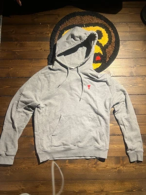 Grå hoodie från Ami Paris - Snygg grå hoodie från Ami Paris med klassisk känguruficka och dragsko i huvan. På bröstet finns Ami:s ikoniska röda hjärtebrodyr. Tillverkad i mjuk bomullsblandning.