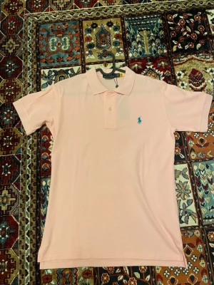 Ljusrosa piké från Ralph Lauren - Snygg ljusrosa pikétröja från Ralph Lauren med klassisk krage och korta ärmar. Tröjan har den ikoniska blå broderade loggan på bröstet och är tillverkad i mjuk bomull. Perfekt för en clean och stilren look.