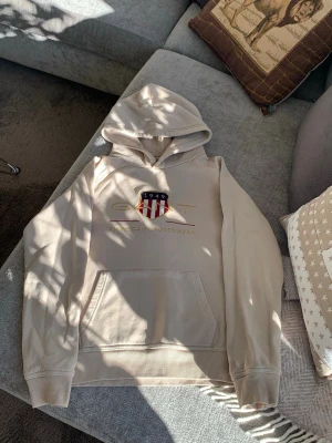 Beige hoodie från GANT med logga - Snygg beige hoodie från GANT med broderad logga och amerikanskt sköldmärke på bröstet. Hoodien har känguruficka framtill, ribbade muddar och huva. Tillverkad i mjukt bomullsmaterial som känns skönt mot huden. Perfekt för en avslappnad och trendig look. Som ny.