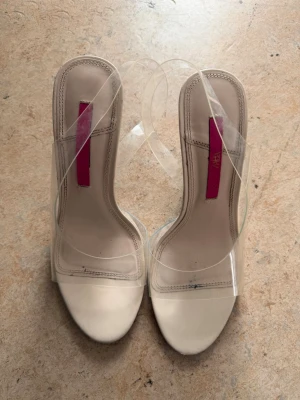 Beige pumps med transparent rem - Säljer ett par stilrena pumps från NA-KD i beige med transparenta plastremmar och smal klack. Skorna har öppen tå och en modern design som ger en trendig look. Perfekta för dig som gillar minimalistisk stil och vill ha något som sticker ut.