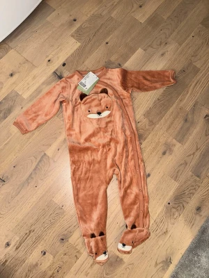 Brun pyjamas med rävansikte 🦊  - Brun pyjamas med rävansikte 🦊 Supermjuk brun pyjamas från Lindex i velour med gulligt rävansikte på bröstet och små öron. Fötterna har broderade rävar och pyjamasen har tryckknappar längs sidan för enkel påklädning. Perfekt för mysiga kvällar hemma.