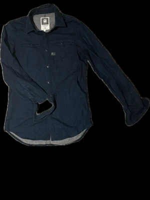 Mörkblå skjorta från G-Star RAW - Snygg mörkblå skjorta från G-Star RAW med klassisk krage, bröstfickor och knappar framtill. Skjortan har lång ärm och är tillverkad i bomull för en bekväm känsla. Perfekt för dig som gillar stilrena och tidlösa plagg.