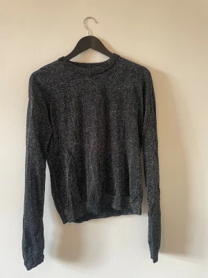 Svart glittrig stickad tröja - En svart stickad tröja med diskret glittereffekt. Tröjan har rund halsringning, långa ärmar och ribbade muddar vid ärmslut och nederkant. Perfekt för dig som vill ha en enkel men ändå lite extra stickad tröja.