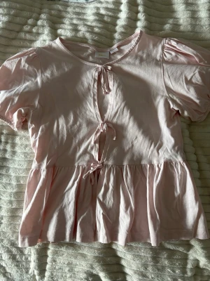 Ljusrosa topp med knyt och puffärm - Supersöt ljusrosa topp med puffärmar och tre knyt framtill. Toppen har en tight passform och volang nedtill. Perfekt nu till sommaren!