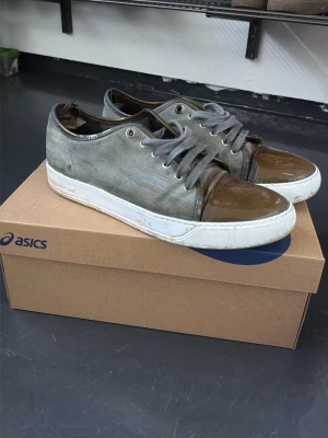 Grå/grön sneakers från Lanvin med lackdetaljer - Snygga grå sneakers från Lanvin med unika bruna lackdetaljer på tån och hälen. Skorna har vita sulor och grå skosnören. Tillverkade i en mix av mocka och lackläder för en cool kontrast. Perfekta för dig som vill sticka ut med din stil. Målade sulor passar 43 