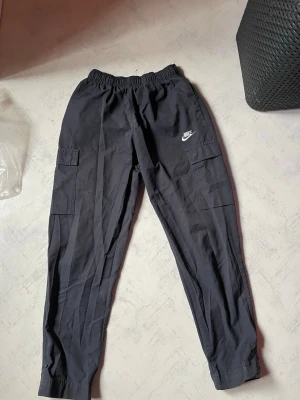 Svarta cargobyxor från Nike - Snygga svarta cargobyxor från Nike med elastisk midja och praktiska sidofickor. Byxorna har en avslappnad passform och resår vid bensluten. Tillverkade i ett lätt och bekvämt material, perfekta för en sportig och casual stil.