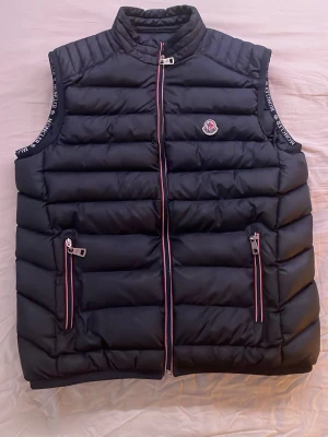 Marinblå dunväst från Moncler - Snygg marinblå dunväst från Moncler med ståkrage och två dragkedjefickor framtill. Västen har Moncler-logga på bröstet och detaljer i rött, vitt och blått längs dragkedjorna. Tillverkad i äkta dun, perfekt för lager på lager under kyliga dagar.