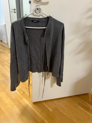 Mörkgrå croppad kofta från bikbok  - Trendig mörkgrå croppad kofta från bikbok  med långa ärmar och knytband framtill. Koftan har en avslappnad passform och ribbstickad kant vid halsen. Perfekt att slänga över en topp för en cool lager-på-lager-look.