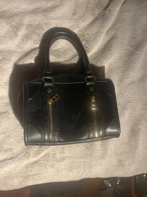 Svart handväska🫶🏼 - Snygg svart handväska i konstläder med två korta handtag och guldfärgade dragkedjor framtill. Väskan har en boxig form och en extra ficka med dragkedja på baksidan. Perfekt för dig som gillar stilrena accessoarer med lite edge.