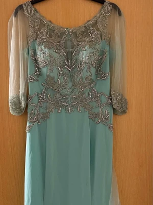 Lång ljusblå klänning med silverbroderi - Elegant långklänning i ljusblått med detaljerat silverbroderi och pärlor över byst, midja och ärmslut. Klänningen har transparenta trekvartsärmar i tunn mesh och en mjuk, flytande kjol. Perfekt för speciella tillfällen där du vill sticka ut.