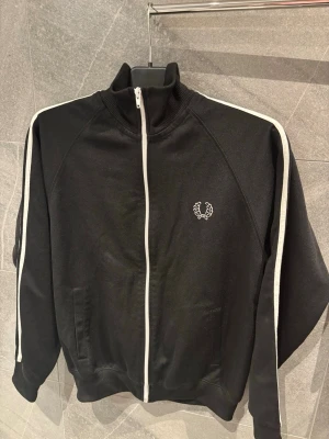 Svart track jacket från Fred Perry - Snygg svart track jacket från Fred Perry med vit dragkedja och vita ränder längs ärmarna. Jackan har ståkrage, två fickor framtill och den klassiska Fred Perry-loggan broderad på bröstet. Perfekt för en sportig och stilren look.