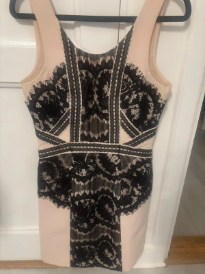 Beige bodyconklänning med svart spets - Snygg beige bodyconklänning med svart spetsdetaljer och mönster framtill. Klänningen har breda axelband och markerad midja som framhäver figuren. Perfekt för dig som gillar en elegant och trendig look.