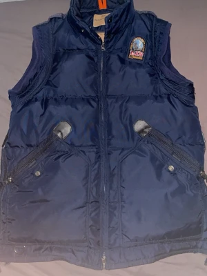 Mörkblå dunväst från Parajumpers - Snygg mörkblå dunväst från Parajumpers med broderad patch på bröstet och två snedställda dragkedjefickor. Västen har hög krage och detaljer i svart läderimitation vid fickorna. Perfekt för lager på lager och streetwear. Priset kan diskuteras 