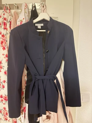 Mörkblå kavaj med knytband - Stilren mörkblå kavaj från Lindex med knytband i midjan och ståkrage. Kavajen har en rak passform, långa ärmar och knäppning framtill. Perfekt för en trendig och elegant look.