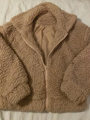 Aprikos  teddyjacka med dragkedja - Mysig aprikos beige  teddyjacka med fluffig yta och hel dragkedja framtill. Jackan har hög krage och är fodrad på insidan för extra komfort. Perfekt för kyliga dagar och ger en avslappnad, trendig look.Perfekt nu till våren.