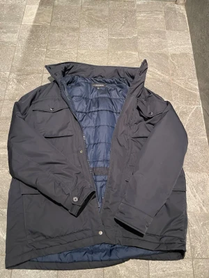 Svart/mörkblå dunjacka från Lindbergh - Snygg svart/mörkblå dunjacka från Lindbergh med quiltad insida och flera praktiska fickor framtill. Jackan har hög krage, dragkedja och tryckknappar samt diskret logga på ärmen. Perfekt för kalla vår dagar och ger även en luftig känsla! 