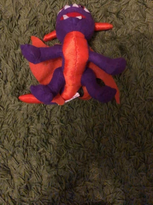 Lila och orange drakmjukisdjur - Säljer ett mjukisdjur i form av en drake med lila kropp och orange detaljer på vingar, taggar och mage. Draken har vita tänder, röda ögon och ett uttrycksfullt ansikte. Tillverkad i mjukt tyg, perfekt som prydnad eller leksak.