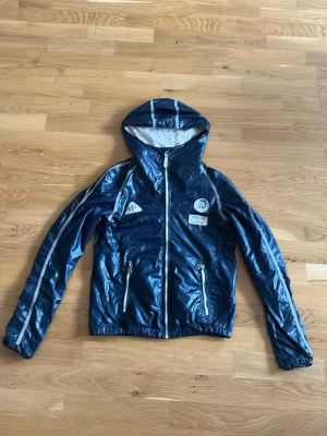 Blå vindjacka med huva och reflexdetaljer - Snygg blå vindjacka med glansig finish, huva och vita reflexdetaljer längs ärmar och huva. Jackan har dragkedja framtill, två fickor med dragkedja och tryckta loggor på bröstet. Perfekt för blåsiga dagar och sportiga outfits. Märket är diesel riktigt skön och snygg perfekt skick 