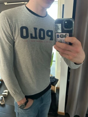 Grå långärmad POLO t-shirt - Snygg grå långärmad t-shirt från Polo Ralph Lauren med stor mörkblå POLO-logga på bröstet och rund halsringning. Tillverkad i mjuk bomull som känns skön mot huden. Perfekt för en avslappnad och stilren look. Några små hål på armbågen syns knappt 