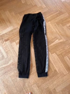 Svarta mesh-mjukisbyxor med logoband - Coola svarta mjukisbyxor med meshmaterial på benen och elastisk midja. Sidorna har vita logoband med text. Perfekta för en avslappnad och sportig look. Ribbad mudd vid benslut och bekväm passform.Skriv gärna för mått! 💕