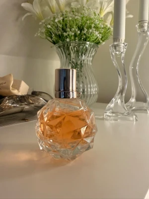 Ari By Ariana Grande EdP 50 ml - Säljer en elegant parfymflaska med diamantformad, transparent glasdesign och silvrigt lock. Innehållet har en varm, persikofärgad ton som ger en exklusiv känsla. Perfekt för dig som vill ha en snygg detalj på sminkbordet.