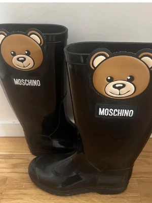 Svarta Moschino stövlar med björn - Säljer ett par svarta stövlar från Moschino med stora björndekorationer framtill och tydlig logga. Stövlarna är gjorda i glansig gummi och har en cool, lekfull design som sticker ut. Perfekta för regniga dagar när du vill ha något extra. Stil 35