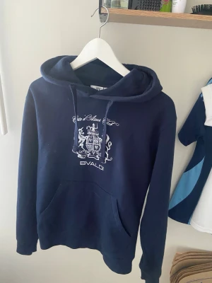 Mörkblå hoodie från Bvaldi - Mörkblå hoodie från Bvaldi med vitt tryck och coolt emblem på bröstet. Klassisk känguruficka framtill och justerbar huva med snören.  Nästan aldrig använd, nypris 899kr