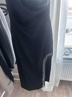 Svart långklänning med strassdetaljer - Elegant svart långklänning med en snygg slits på sidan dekorerad med glittrande strass. Klänningen har en figurnära passform och en stilren design som ger en lyxig känsla.