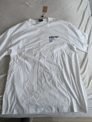 Vit Stüssy t-shirt med jordgubbstryck - Vit t-shirt från Stüssy med blå logga och text på bröstet samt en stor jordgubbe och logga på ryggen. Klassisk passform och rund hals. Perfekt för dig som gillar streetwear och vill sticka ut lite extra.