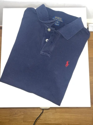 Marinblå pikétröja från Polo Ralph Lauren - Klassisk marinblå pikétröja från Polo Ralph Lauren med röd broderad logga på bröstet. Tröjan har krage, knappar framtill och är tillverkad i bomull för en bekväm känsla. Perfekt till jeans eller shorts för en clean look.