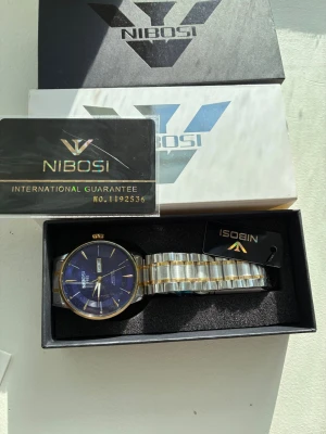 NIBOSI klocka blå  silver/guld - Stilren klocka från NIBOSI med blå urtavla och datumvisning. Armbandet är i silverfärgad metall med gulddetaljer. Kommer i originalförpackning. Perfekt accessoar för dig som vill ha en klassisk men modern look. Aldrig änväd 