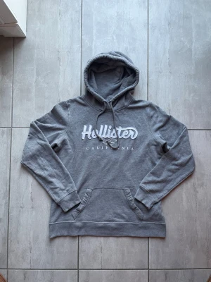 Grå hoodie från Hollister - Snygg grå hoodie från Hollister med stor vit logga och texten 'California' på bröstet. Klassisk känguruficka framtill, ribbade muddar och justerbar dragsko i huvan. Perfekt för chill dagar och enkel att matcha med jeans eller mjukis. Passar strl: S